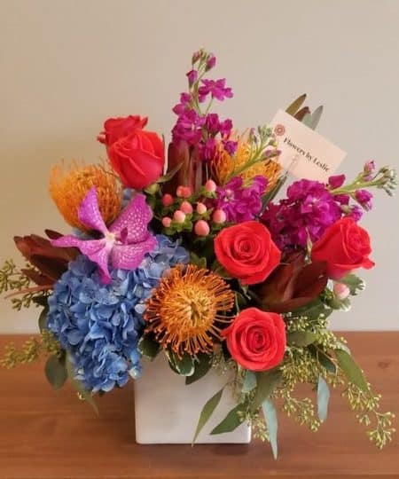 A vibrant grouping of roses, pin cushion protea, hyperium and hydrangea in reds, oranges and blues. All the colors of a beautiful autumn sunset...